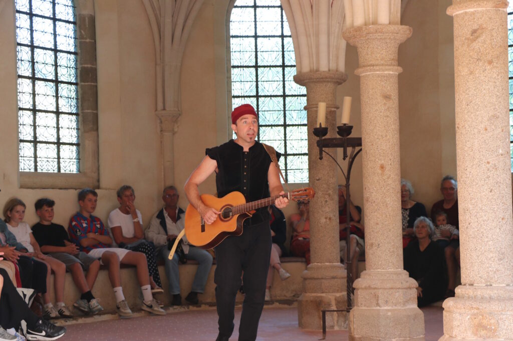 Un troubadour chante dans la salle capitulaire devant le public assis sur les bans tout autour de l'abside.