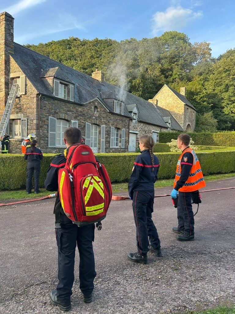 Pompiers de Hambye en exercice devant l'ancienne maison du gardien de l'abbaye de Hambye. Trois pompiers observant leurs collègues et la fumée sortant d'une fenêtre.