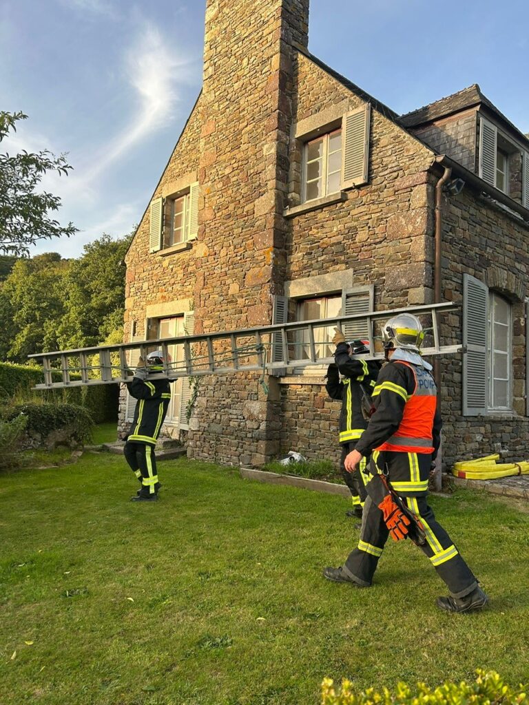 Pompiers de Hambye en exercice devant l'ancienne maison du gardien de l'abbaye de Hambye. Deux pompiers portant une echelle.