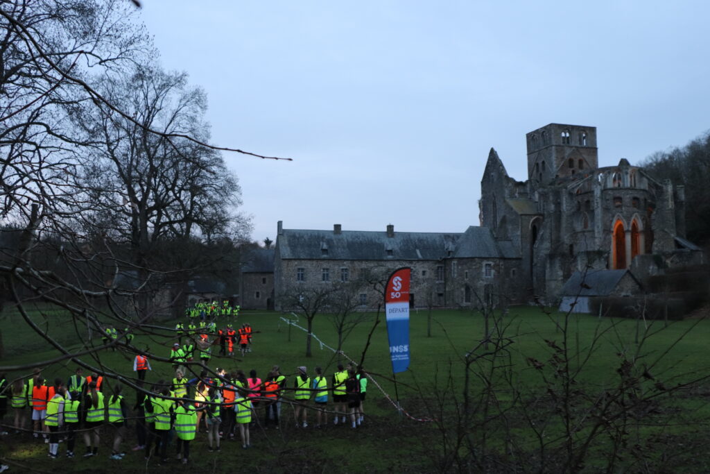 Départ du trail semi nocturne dans les jardins monastiques de l'abbaye de Hambye. La nuit tombe, des élèves en tenue de sport et en gilet jaune se trouvent au premier plan ainsi qu'une flamme mentionnant l'UNSS, organisateur de l'événement. Les bâtiments monastiques et le chevet de l'église abbatiale sont en arrière plan.