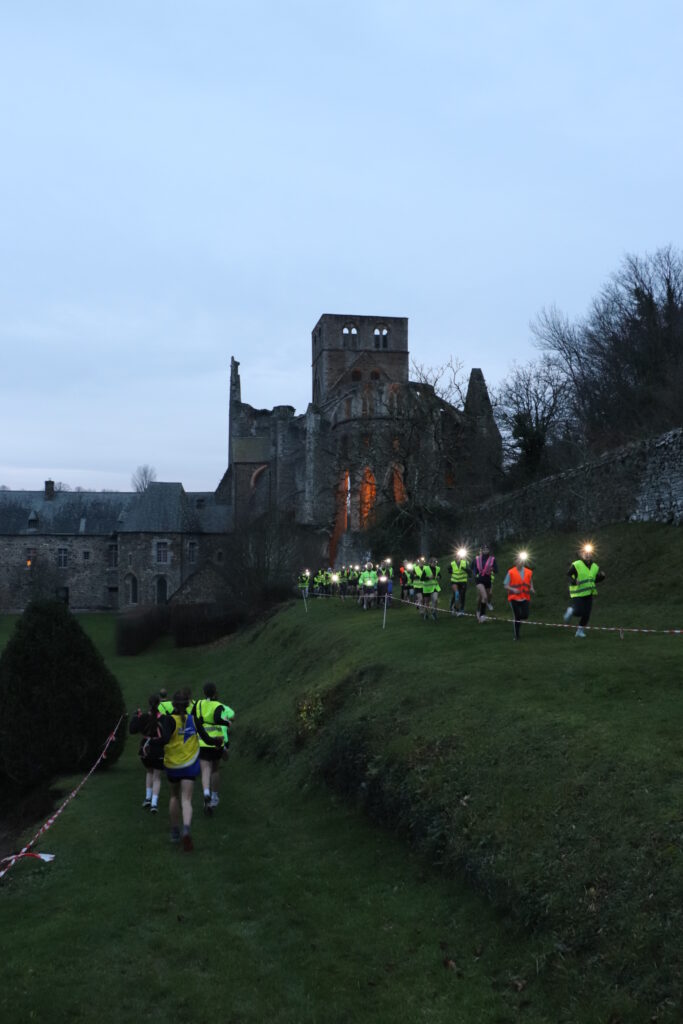 Les élèves participant au trail semi-nocturne au pied du mur d'enceinte dans les jardins monastiques. Ils ont des lampes frontales allumées sur la tête et des gilets jaunes. Ils courent en file indienne.