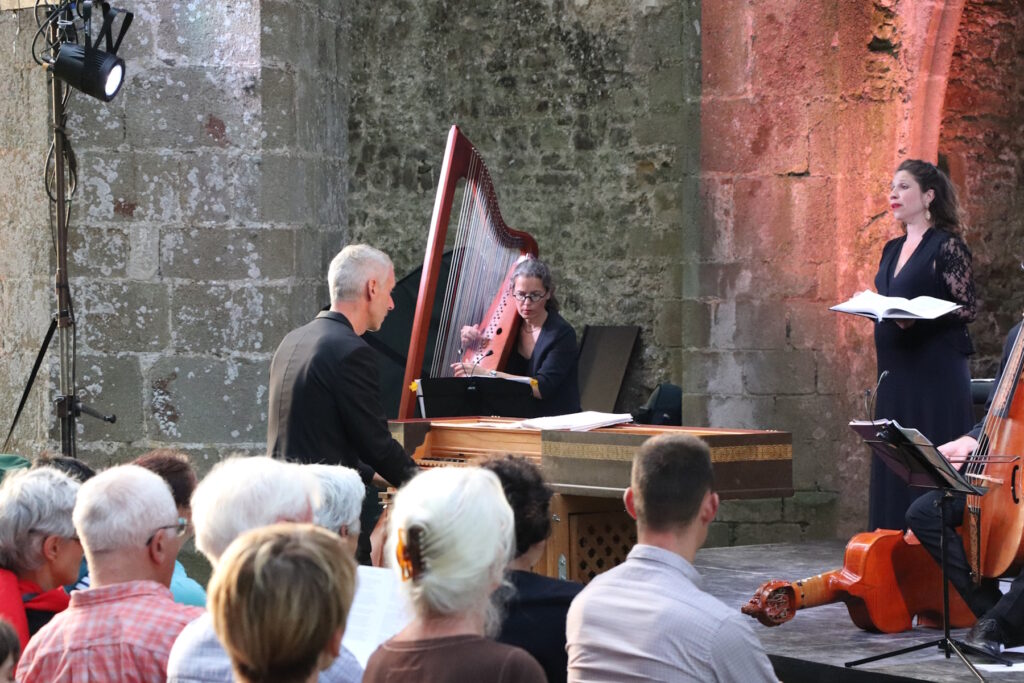 Un musicien est assis derrière un clavecin. On voir également une harpiste et une chanteuse. Le concert se déroule le soir dans l'église abbatiale.