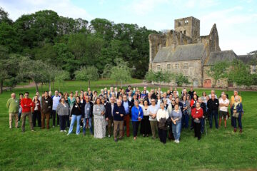 90 partenaires du festival Pierres en Lumières dans la Manche posent dans le verger de l'abbaye, avec l'église abbatiale et l'aile des convers en décor de fond.