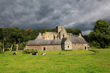L'abbaye de Hambye vue depuis la route de Percy avec au premier plan les bâtiments agricoles. Le ciel est rempli de nuages gris
